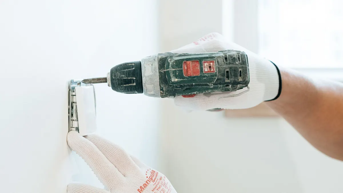 Gloved hands using a drill to install a power outlet on a wall, focusing on precision and safety.