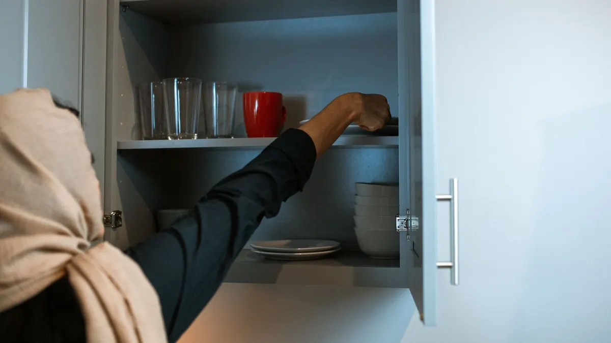 A woman arranges dishes in a kitchen cabinet, reaching out to organize tableware.