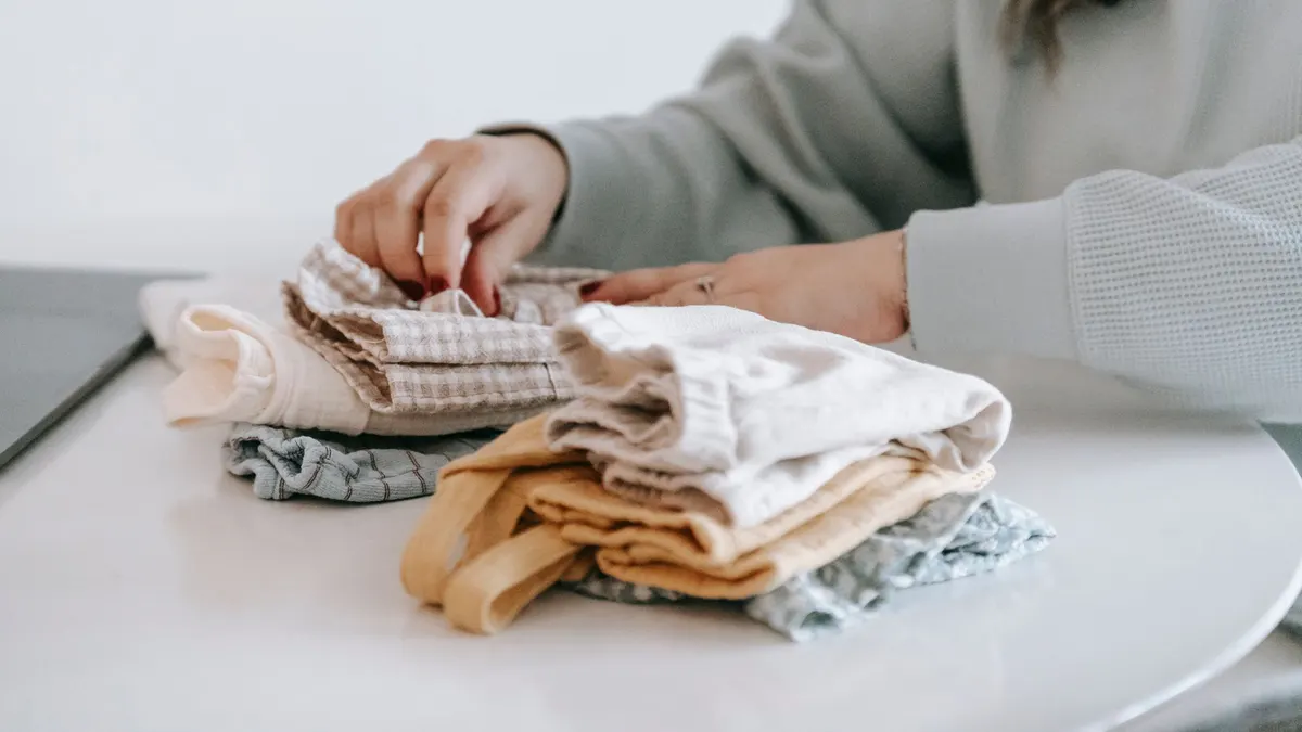 Crop faceless female in casual clothes folded colorful baby garments while sitting at white table in room on white background