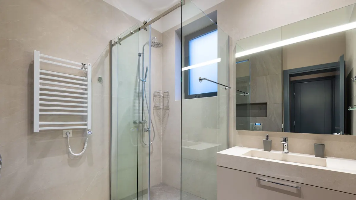 Shower cabin with glass walls near mirror hanging on wall above sink in modern minimalist styled bathroom
