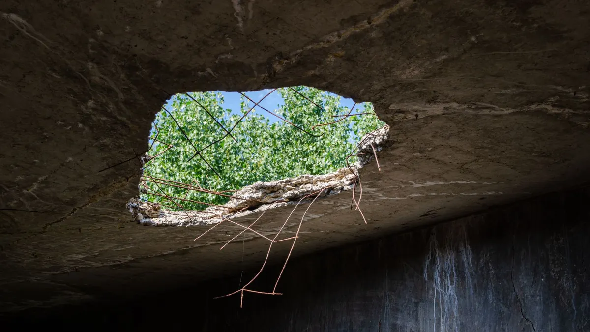 A large hole in a concrete structure provides a view of greenery and blue sky.