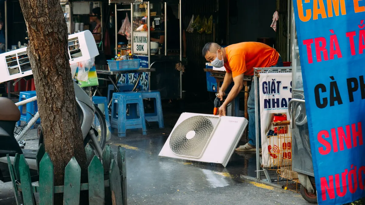 A man cleans a fan on a vibrant urban street with market stalls.