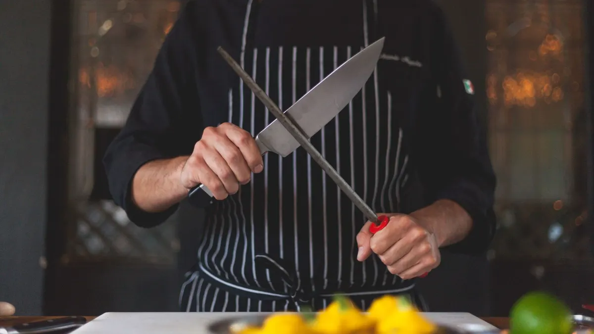 Chef sharpening a knife with a honing rod in a professional kitchen setting, focusing on precision and culinary skill.