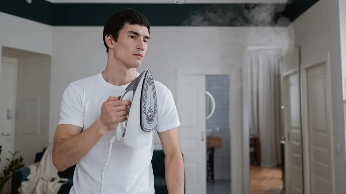 A man uses a steam iron on a white shirt indoors, highlighting daily household chores.