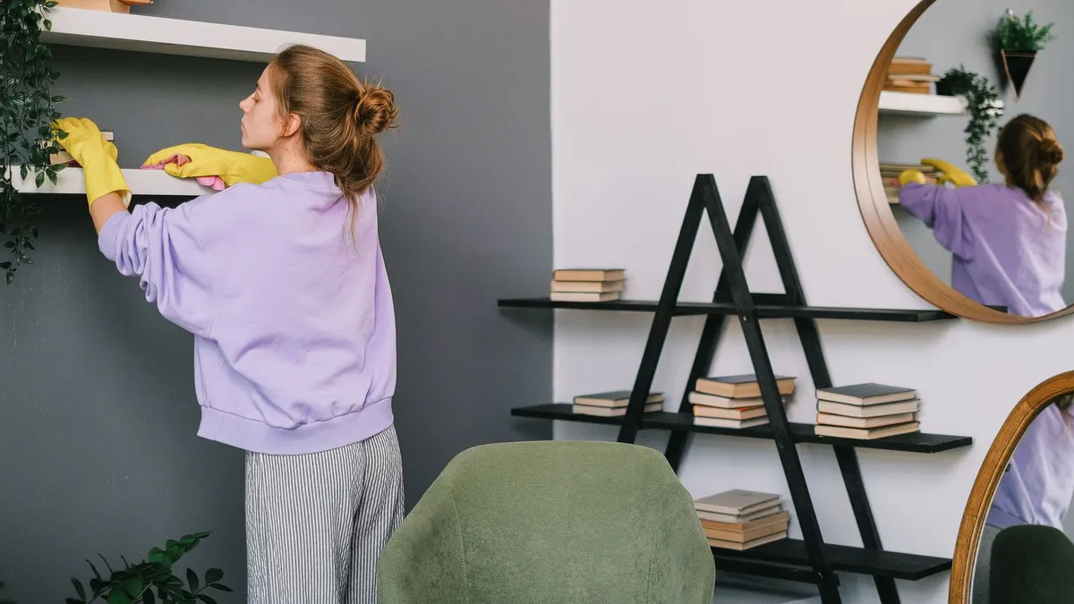 A woman cleans shelves in a stylish living room with mirrors and decor.