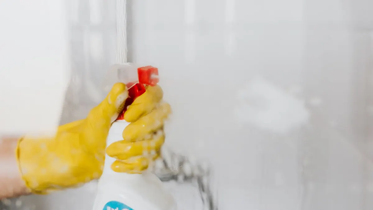 Crop unrecognizable person in yellow latex protective glove spraying detergent from bottle with plastic dispenser at glass of shower cabin