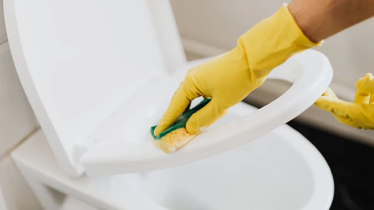 From above of crop faceless person in yellow rubber cleaning gloves cleaning plastic toilet seat properly with sponge in restroom