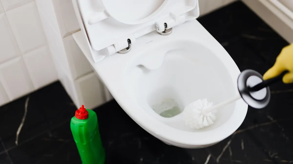 From above of crop anonymous person in rubber gloves using liquid toilet sanitizer and brush to clean toilet bowl in bathroom