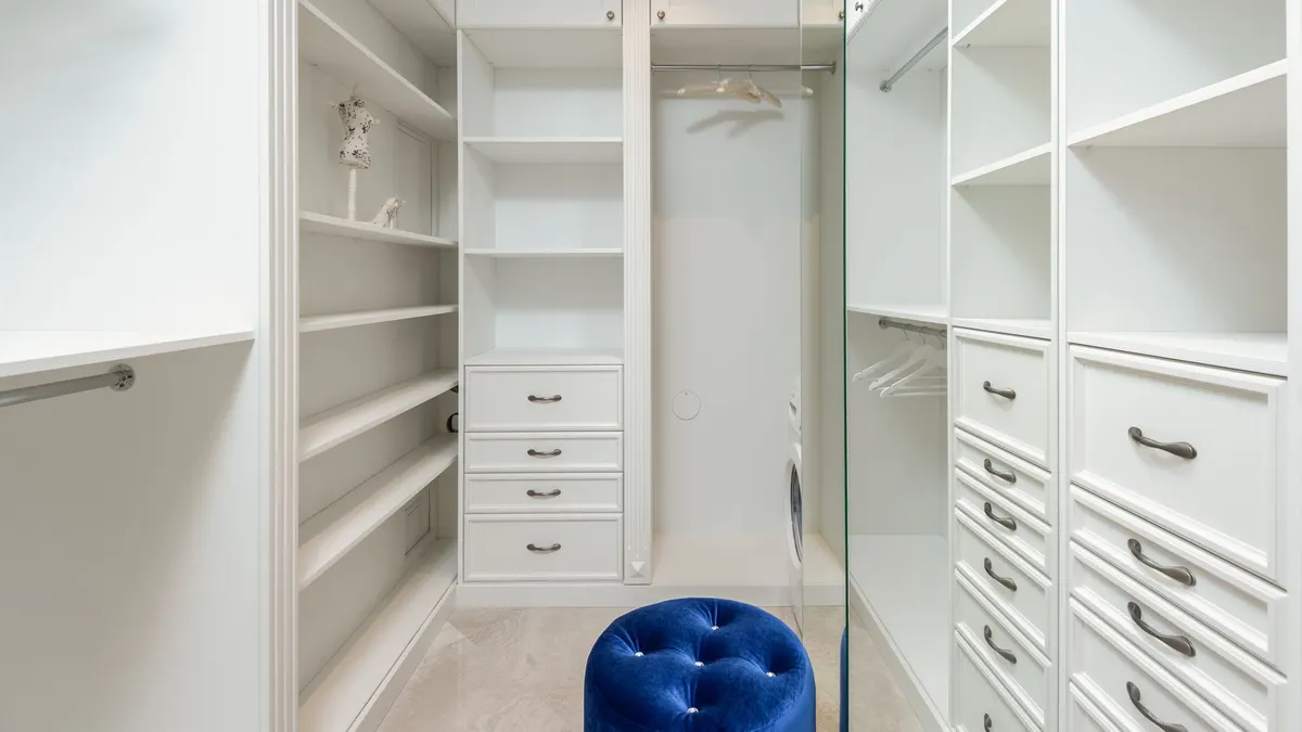 Soft blue velour pouf in white built in closet with empty shelves and racks in classic styled apartment