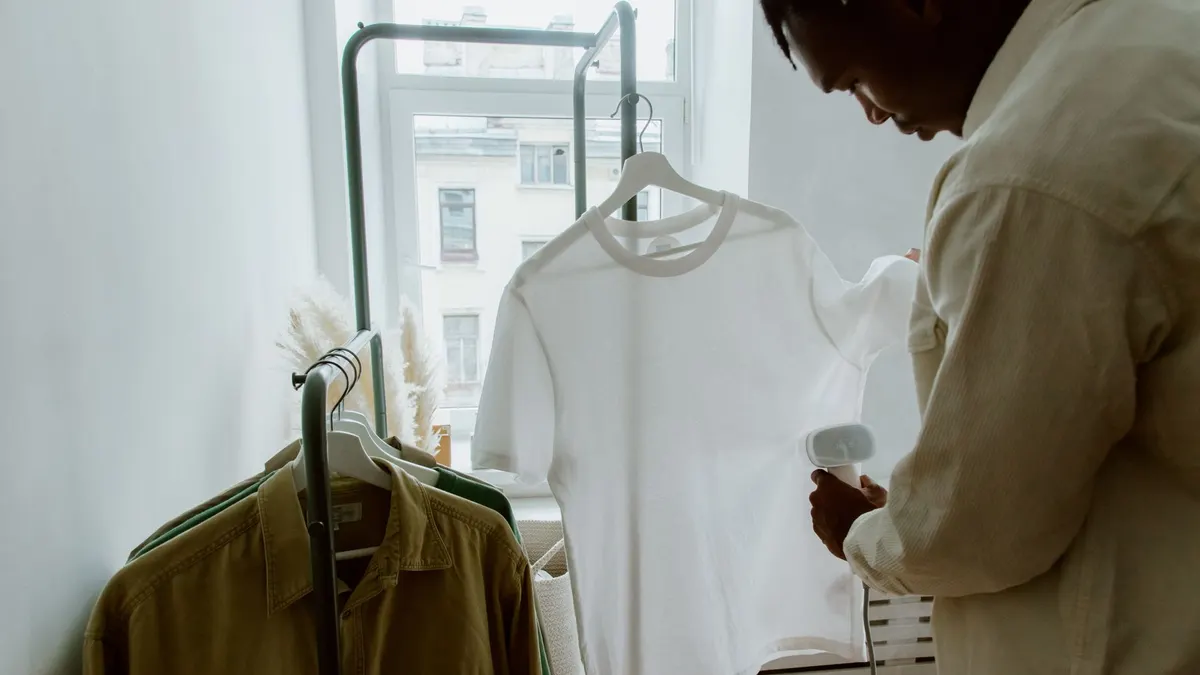 A man uses a steam iron on a white shirt hanging from a clothing rack indoors.
