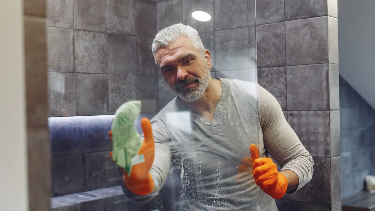 Grey haired male with beard in orange rubber gloves attentively cleaning shower glass in bathroom